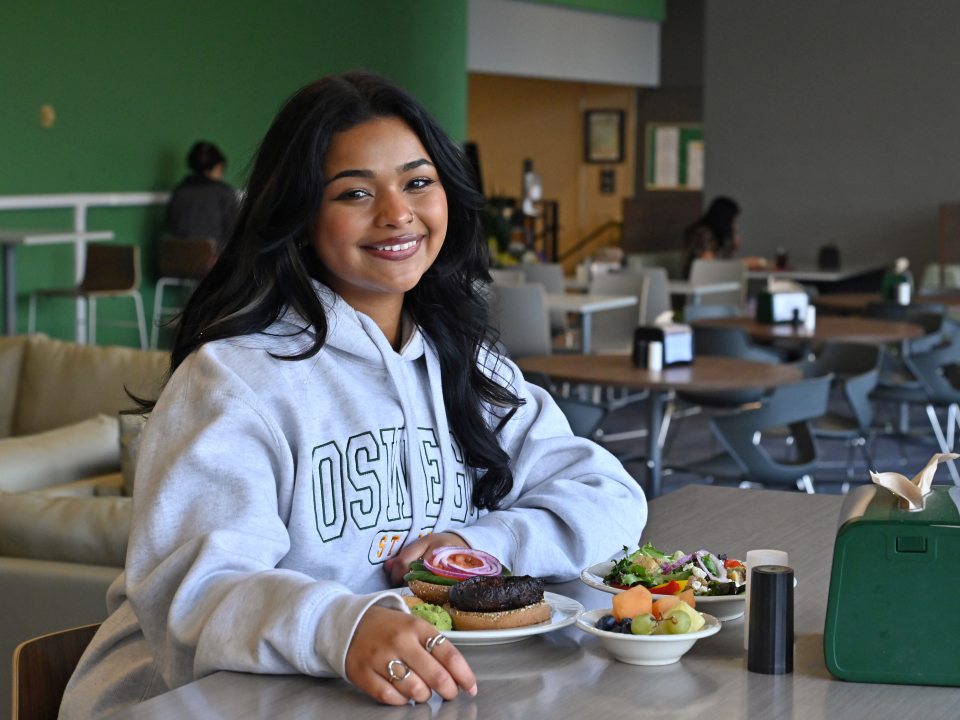 A student wearing an Oswego sweatshirt smiles while seated at a dining hall table with a burger, salad and fresh fruit.