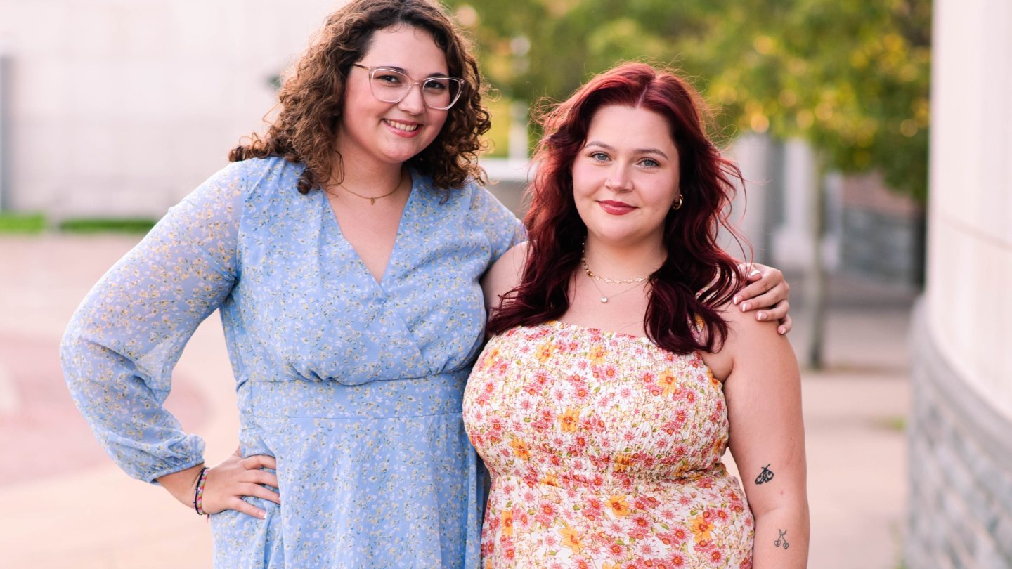 Two Office Staff members dressed up, smiling and looking into the camera.