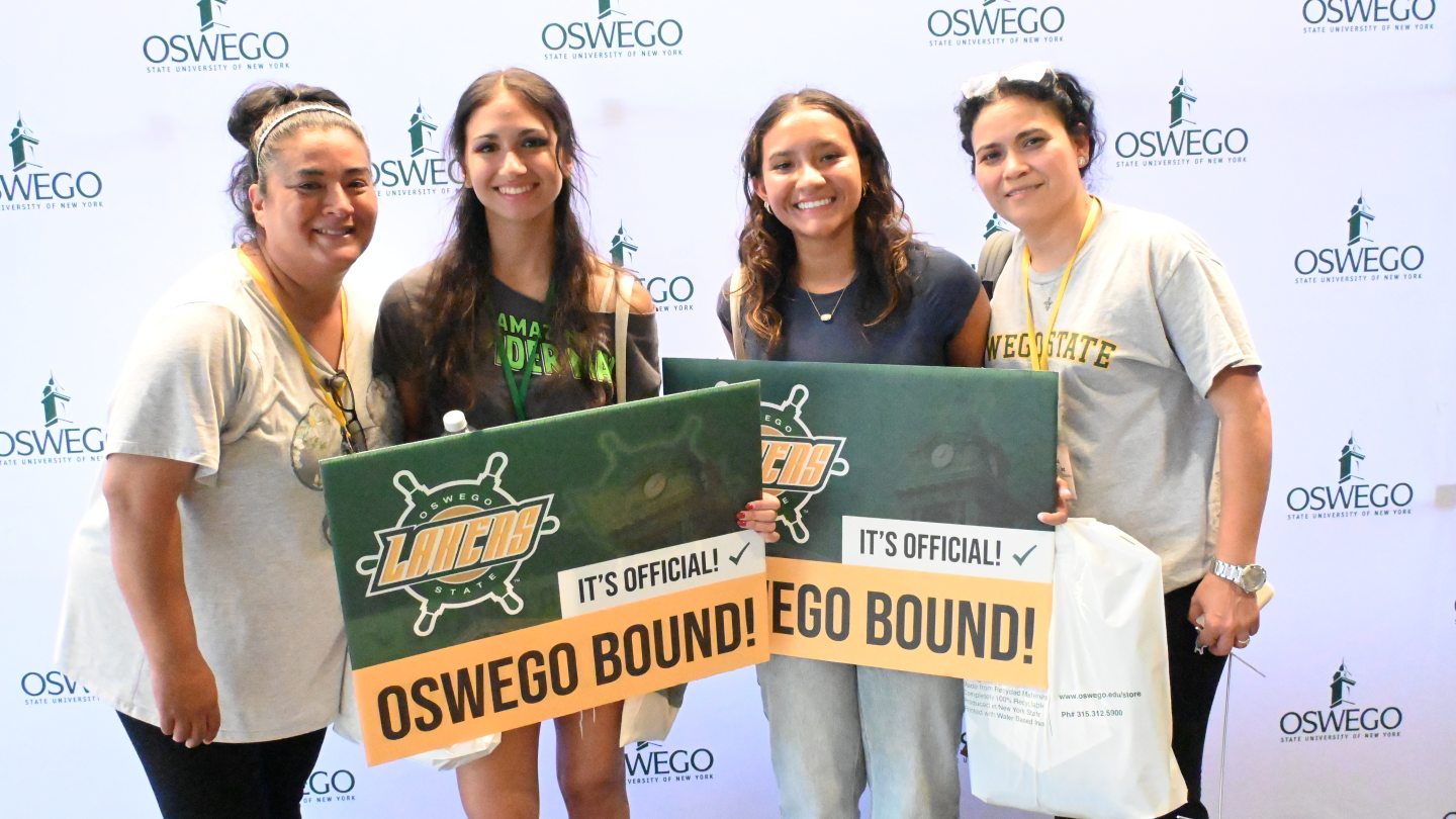 Two student and their supporters holding "Oswego Bound!" signs with a photo wall in the background