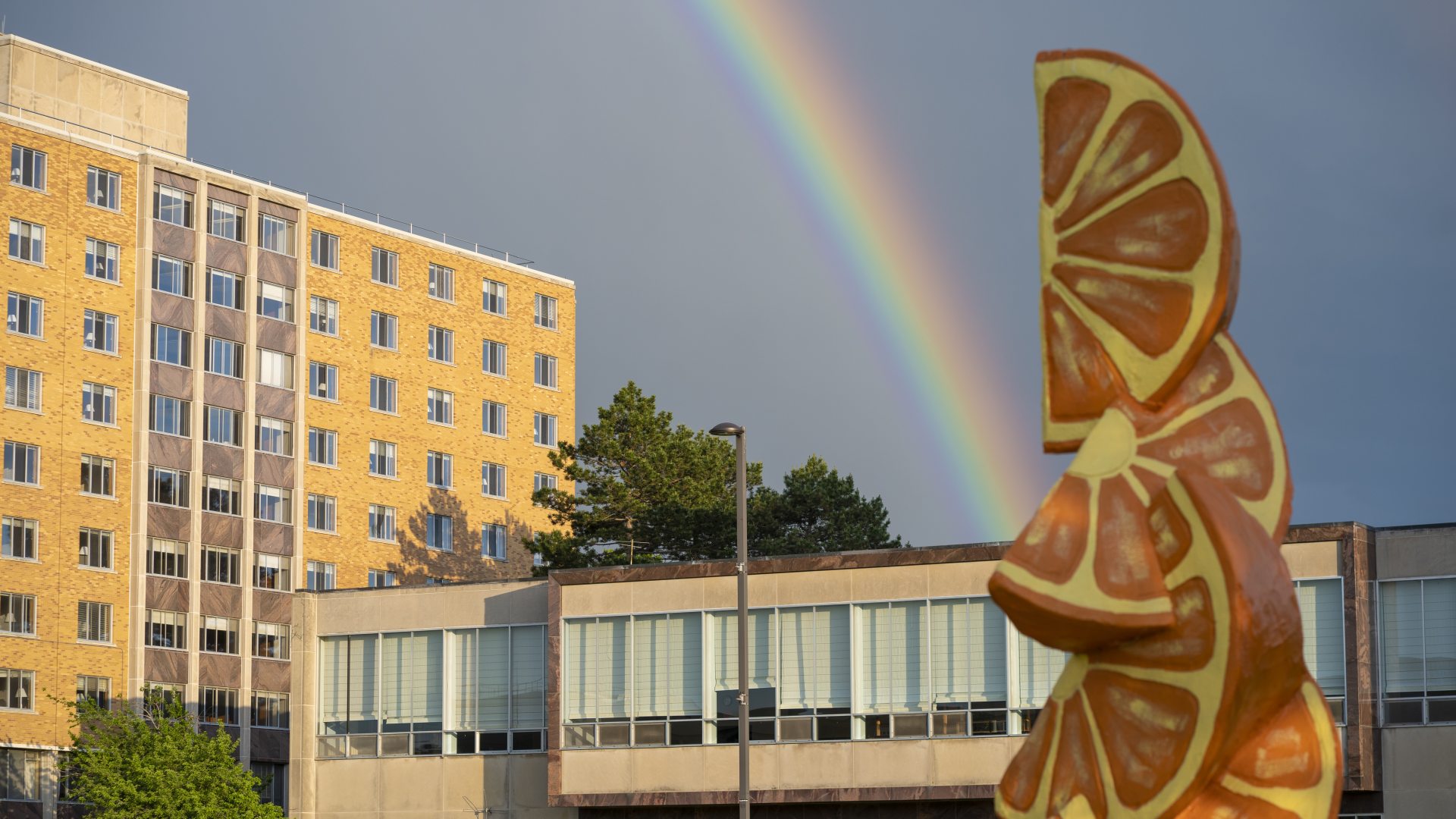 A photo of a rainbow stretching over Cooper Dining Hall, with an orange statue  from the quad in the forefront.