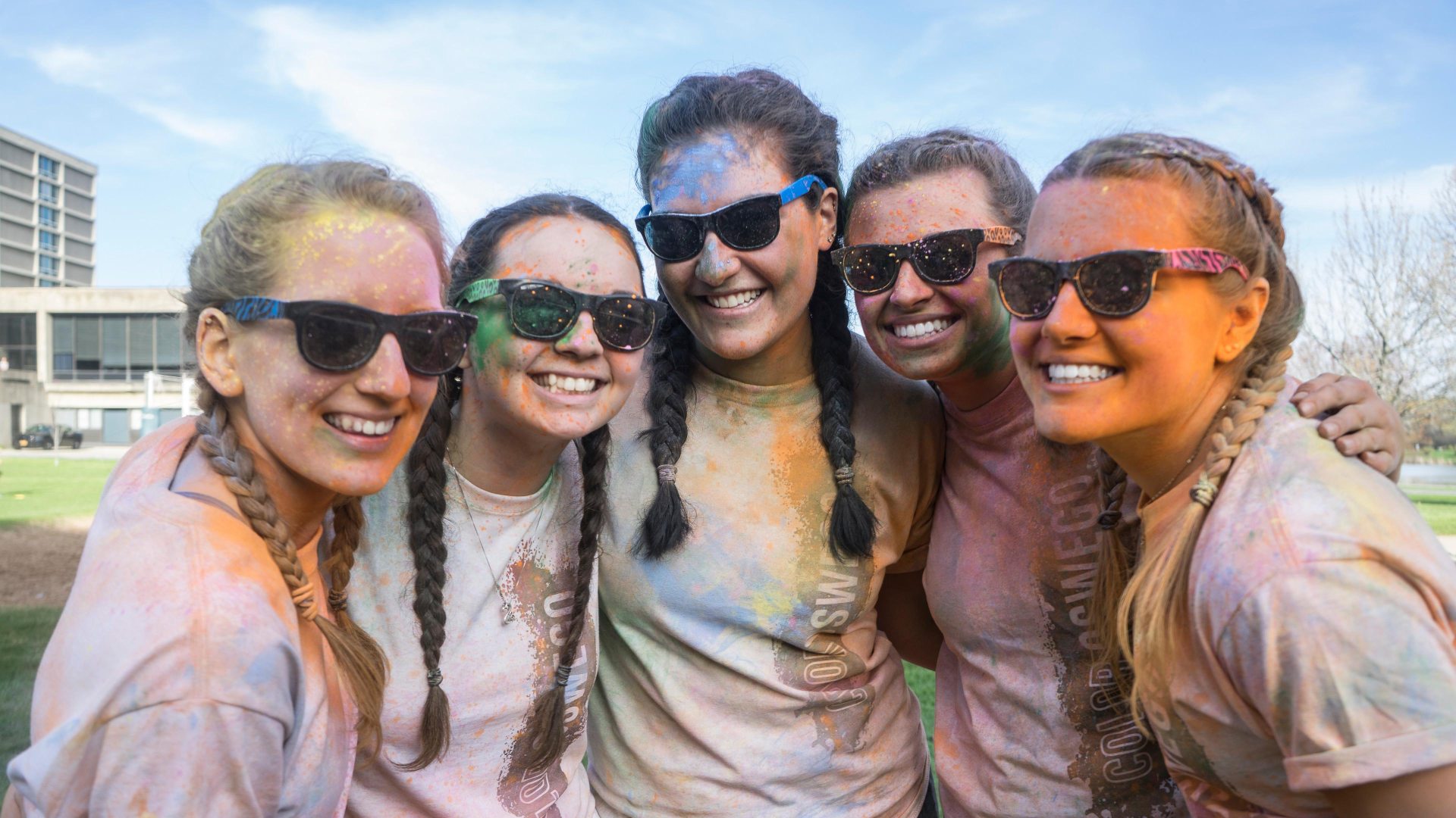 A group of women wearing sunglasses, shorts, and white T-shirts with colored powder on them from doing a Color Run