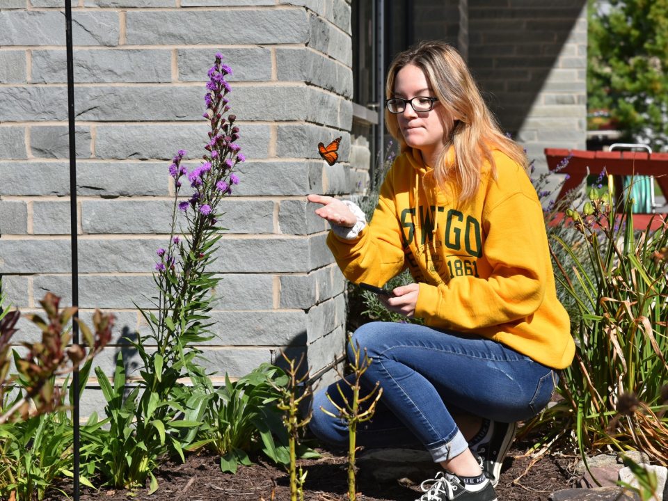 Student at Rice Creek Field Station kneeling in a garden holding her hand under a Monarch Butterfly