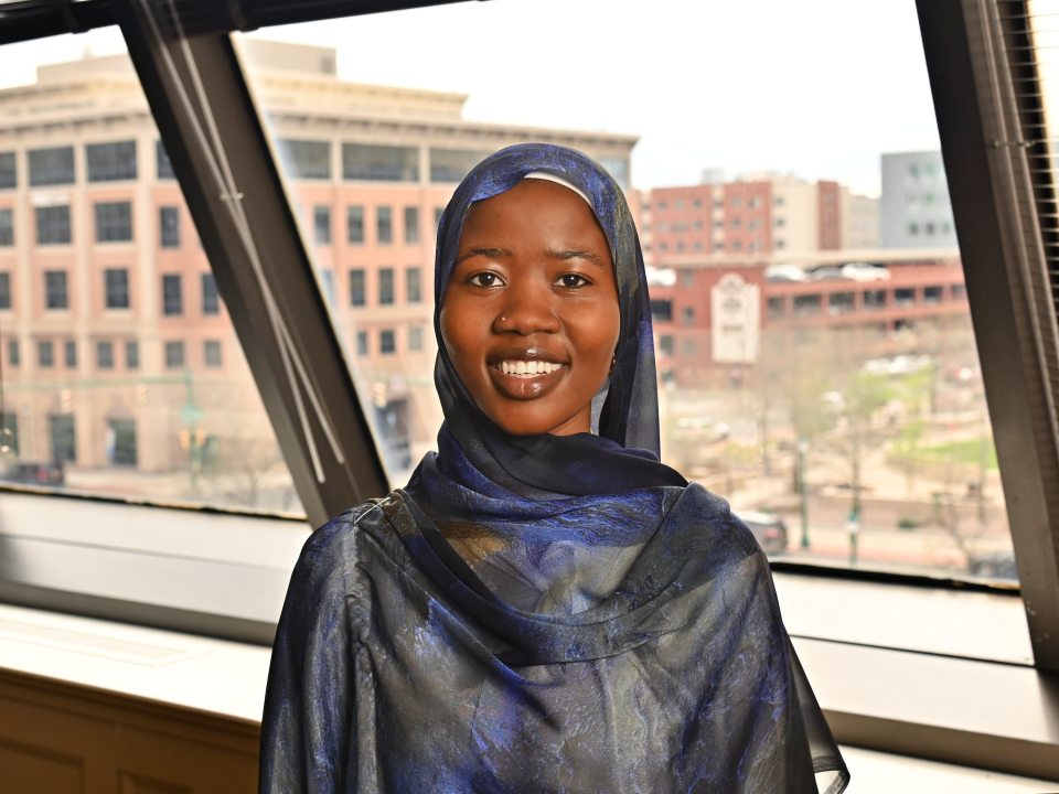 Adult student wearing a blue patterned hijab smiles while posing inside SUNY Oswego at Syracuse, with downtown Syracuse buildings visible through large windows in the background.