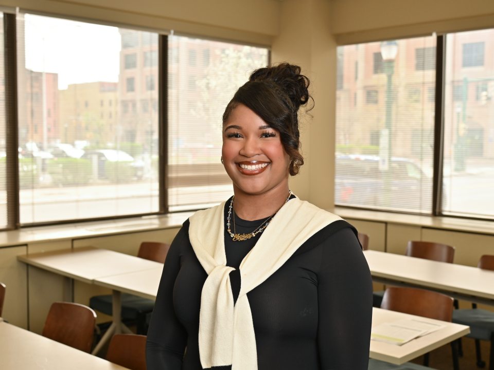 Adult student smiling and posing in a classroom at SUNY Oswego at Syracuse, with large windows and downtown Syracuse buildings visible in the background.