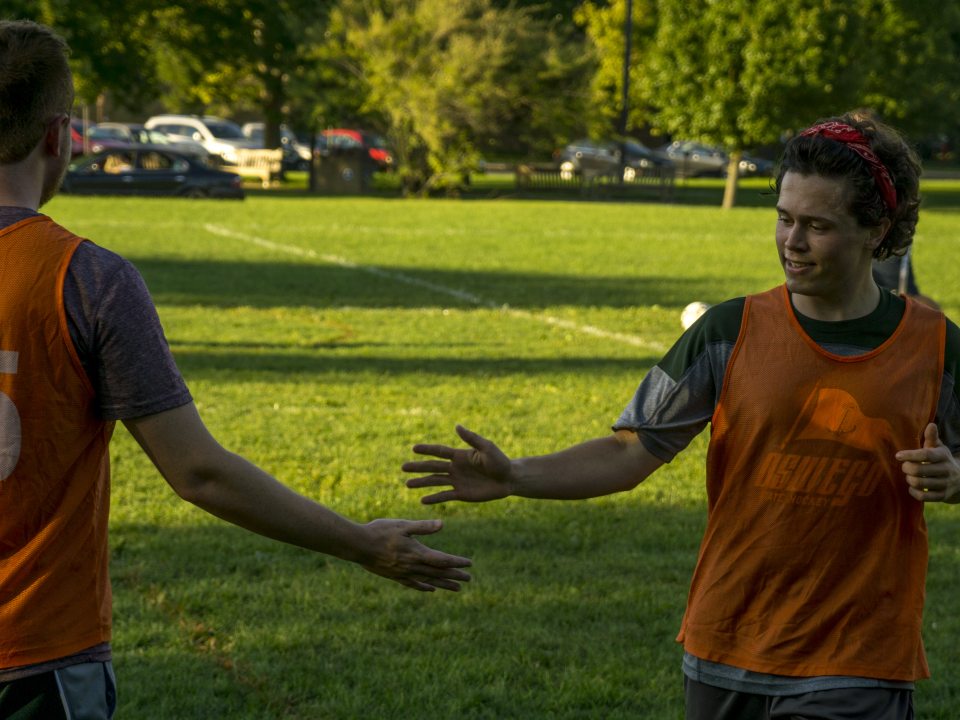 Two people wearing orange jerseys on a playing field, slapping hands as they pass each other 