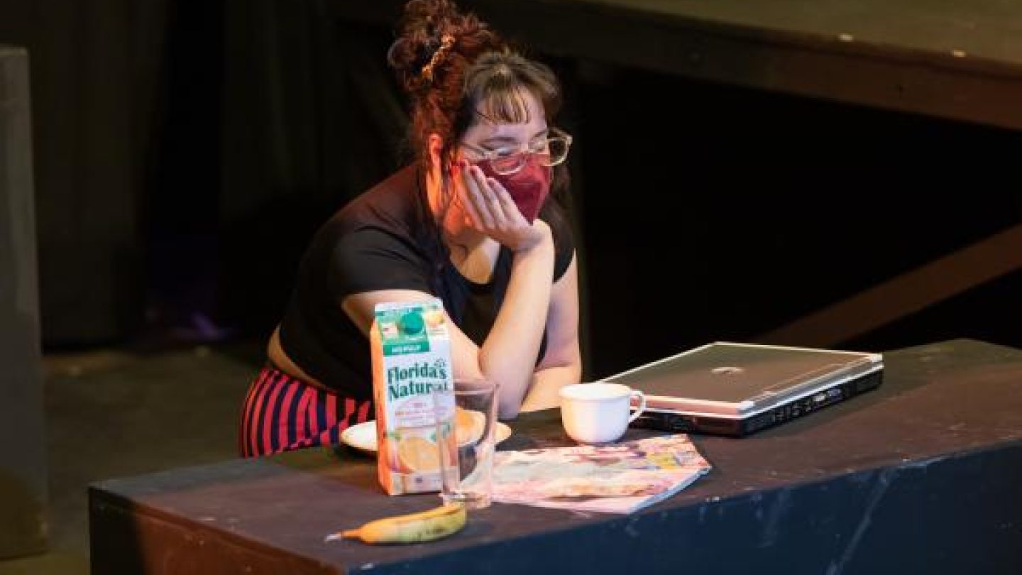 A female student sits at a table with a laptop, banana, tea cup, and a carton of orange juice