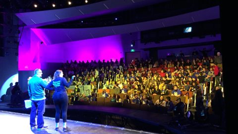 Two people standing on Waterman stage addressing a full house audience
