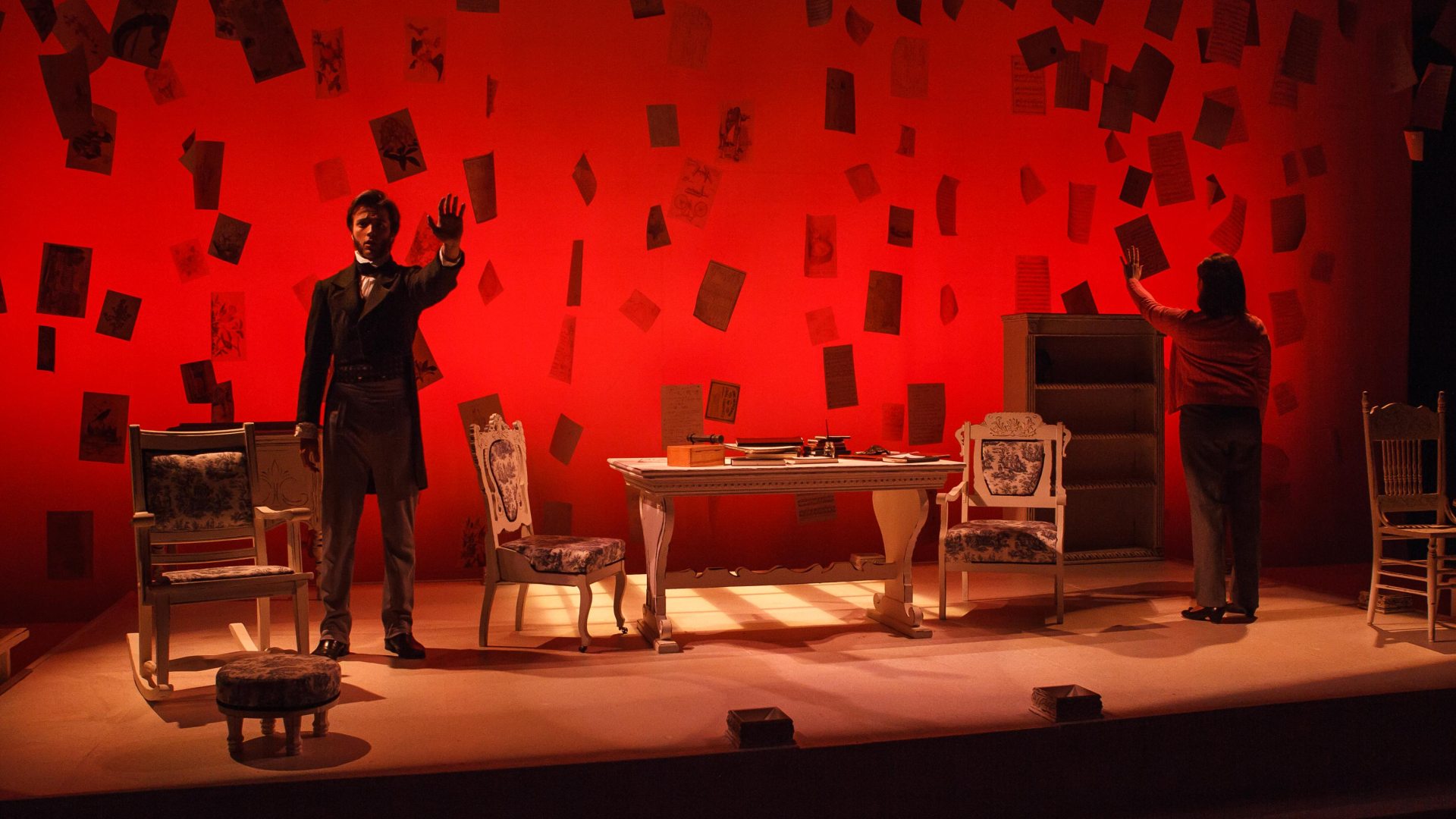 two actors on stage acting out a scene; one actor on the left is holding out his hand and the actress on the right has her back to the audience but is holding out her hand