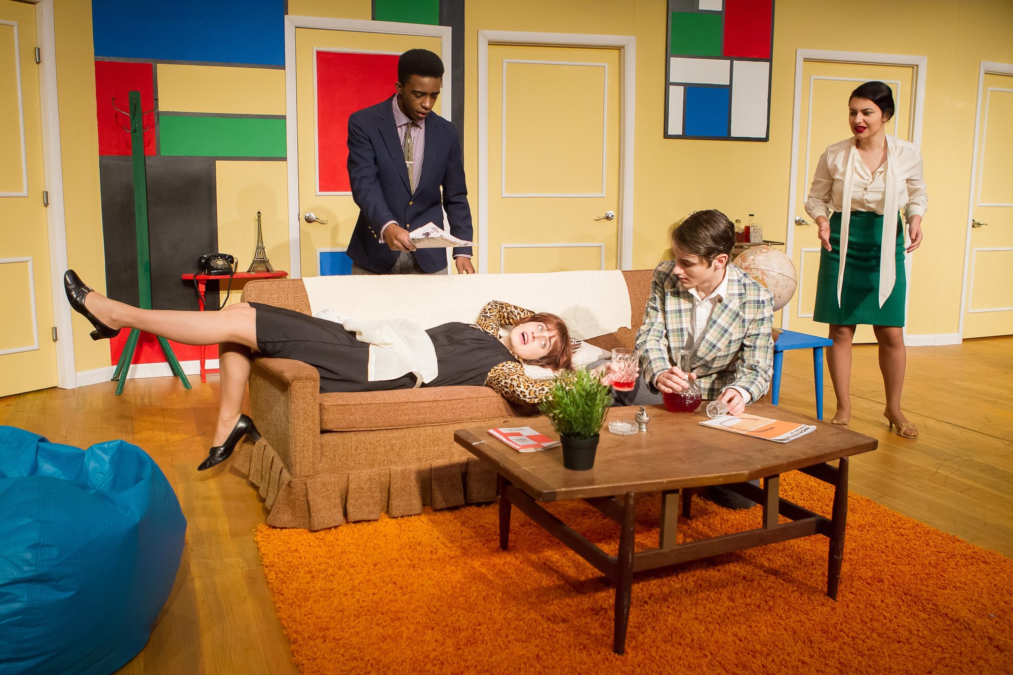 student actors on stage in a colorful living room acting out a scene, one actress is laying on the couch in the middle with another actress sitting next to her. Two actors are standing near the couch. 