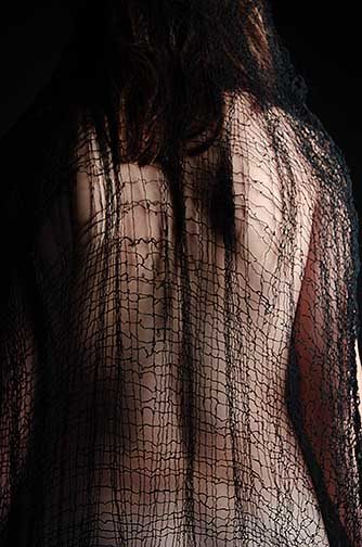 A portrait framed photo of the back of a woman, with the tom of her head and the base of her back cropped. Sprawled across her back is that same black shawl which goes from the length of her head to the bottom of her back, covering both arms as well. 