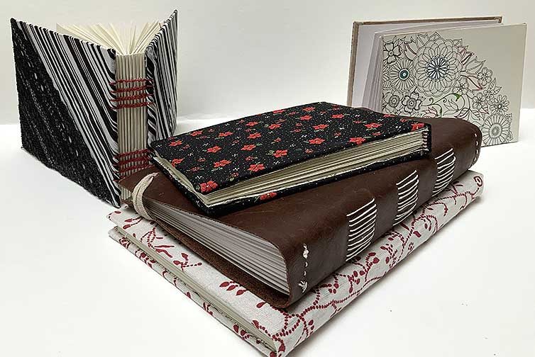 A grouping of five different, blank books that were handmade and bound. A Coptic book with red string and a black and white covering, an accordian book with a fun coloring page as it’s covering, a perfect bound book with a hard spine covered in a floral fabric, and two soft perfect bound books. One covered in leather and the other covered in a red and white fabric.