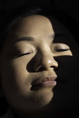 This image is of a tightly framed portrait of a girl with harsh shadows. Darkness surrounds the face, framing the subject matter and pushing it forward. Her face has no expression on it as her eyes and lips are gently closed with her skin showing much detail and shadow. There is a hard shadow vertically across her face with a rectangle framing her right eye. The shadow continues around the entirety of the face closing out all surroundings.