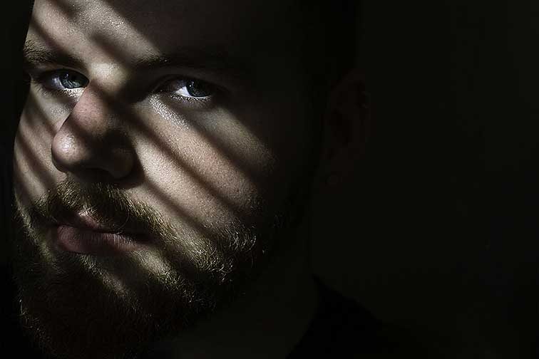 This photograph shows the face of a male model with multiple hard vertical lines of light and shadow across the face. The skin shows much detail such as pores and facial hair. His blue eyes are prominent as the light illuminates the color. As the subjects face shifts to the right, his eyes are looking into the camera with a straight face. The subject is on the left side of the photograph with negative space surrounding his face and right side.