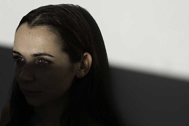 This image is of a female subject against a white wall with a diagonal line cutting across the face. This line separates the top and bottom half of the photo with light and shadow. This shadow starts below the eyes and above the nose while being continuous from left to right. The subject is on the left side of the photograph and looking into the light source instead of the camera. The photograph is cropped from her neck and up with negative space on the right side and above her head.