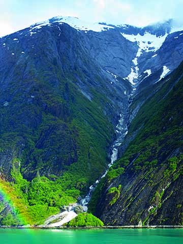 Vertical photograph of a mountainside with a stream running down the middle that has ice and snow in it. The clouds are obscuring the top of the mountain, leading down to dark grey shall on the mountainside. The shall transitions into dark green foliage. The bottom is washed in sunlight, making the foliage a brilliant medium green-yellow. There is a small rainbow in the bottom left corner over the teal water.
