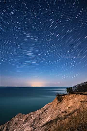 A long exposure photograph showing the movement of stars in the night sky above the Bluffs and Lake Ontario in Sterling, New York