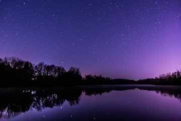 A photograph of a glassy, calm river reflecting a purple night sky and stars above it in Fulton, New York.