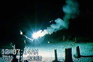 A VHS Image of a man outdoors in the dark, he is holding a blue sparkler and there is a lot of smoke surrounding it.