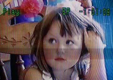 A VHS image of a little girl sitting at a table, she is wearing a birthday hat and is looking off to the left. She is playing with her hair and looks unamused. 