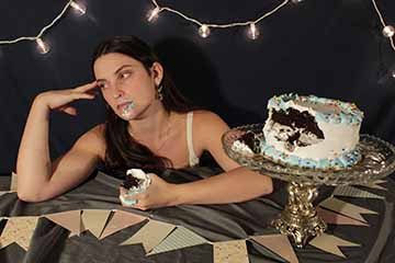 This a photo of a dark haired white woman sitting behind a table with a cake on a stand next to her. She has a chunk of the cake in her hand and she is looking to the side away from the cake. Her expression is irritated. On the table is a grey table cloth and pink party decorations. Behind her is a dark blue background with string lights.