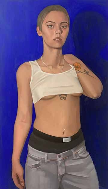 This is a portrait of white woman with light brown hair and a very short haircut. She has one hand on her shoulder and the other down by her side. She is wearing large jeans, black boxer underwear pulled up. She has a white tank top cut short to show the under part of her breasts. She has a butterfly tattoo on her sternum and flowers on her forearm. The background is bright blue. 