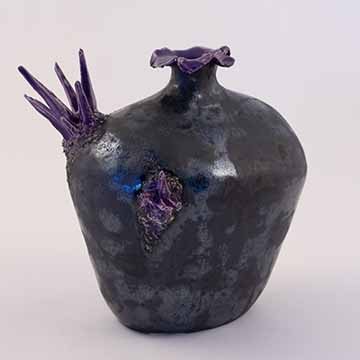 Image a: A black vase with purple crystal forms growing out of it. The vase is narrow at the bottom, wide at the shoulder, and closes to a very thin neck with an undulating rim. A grouping of crystals points out of the left shoulder of the vase. A second grouping points toward the camera and is slightly down and right of the first group. The surface is mottled matte and metallic in the black area. The crystals and top of the rim are a glossy purple.