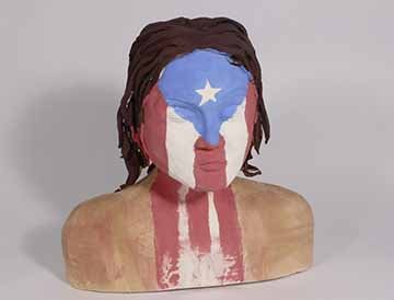 A ceramic bust of a girl from the shoulders up. Her eyes are closed and has brown wavy hair. The girl has bare shoulders and the Puerto Rican flag painted over her face to the bottom of the bust. 