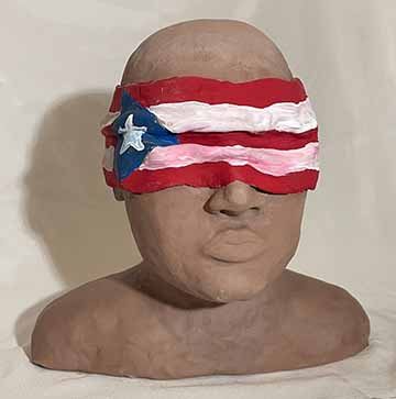 A bust of a woman from the top of her shoulders. She is made with clay and is not painted with the exception of a bandana made of clay made to wrap around her eyes and head. She does not have hair or any distinguishing features.