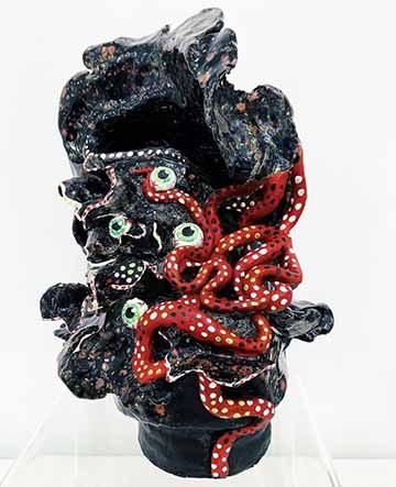 This ceramics piece contains gestural petal-like edges, and on the right side, there are red, white and yellow tentacle-like shapes that connect the top to the bottom half. There are spheres intertwined within the tentacles, with painted eye shapes on them. The body of the form is black with red and yellow speckles, and there are green tones within the edges as well. The form serves as a vase-like decor, because it is able to physically hold things inside of it.