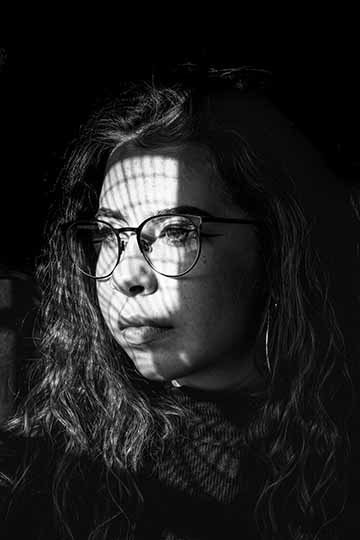 This is black and white photograph, capturing a three quarter view of a woman&#039;s face. She is looking off into a window, a unique shadow-casting lighting sits across her face. 