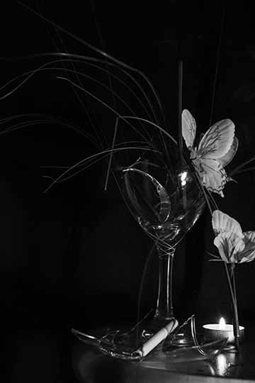 “Broke Up” is part of a digital photograph series titled “The Quarantine Studies”, that was created in the height of the COVID-19 pandemic. The black and white image consists of a broken wine glass with the broken pieces in and around the glass. There is a line of plants and flowers appearing to come out of the broken glass, while a lit cigarette and burning candle can be seen at the bottom of the frame. Heavy contrast and dark blacks are used throughout the photo, giving it a dramatic feel.