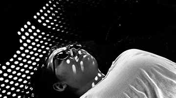 This is a black and white photo of a person laying on the ground underneath a park bench. The holes in the bench creates an interesting pattern on her face as the sunlight hits the bench.