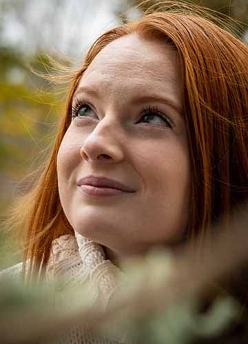 The photography is a colored, up-close portrait shot of a female subject looking up to the left into the abyss. The female has beautiful red hair complementing a white turtleneck. There is a tree branch with green leaves cutting across the image at the lower left rising the lower right side in a diagonal motion. 