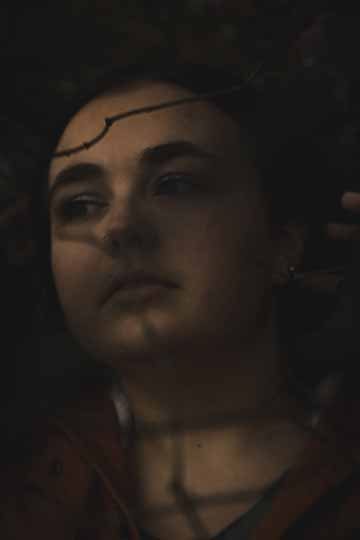 The photograph is a portrait shot of a female subject that is looking away from the camera, brushing hair behind her ear standing behind tiny tree branches that lay in front of her face. 