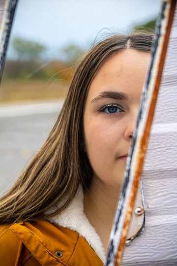 The is a colored close up portrait photograph of a female subject. Only half of the female model’s face is being shown in the reflection of an old rusty van mirror.