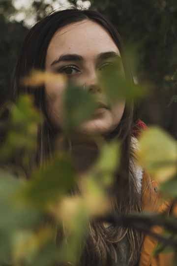 The photograph is a close up portrait shot of a female hidden behind leaves from a large tree branch. 