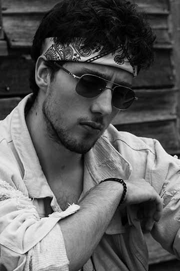 The photograph is a close up black and white photograph of a male, wearing old school aviators, a white head- hand and a destroyed jean jacket looking down at his feet but far off into the abyss.