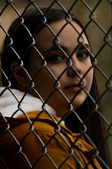 The photograph is a portrait shot of a female subject looking directly into the camera behind an old, wire fence. 