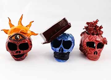 ​​​​​​​This piece includes three ceramic skulls. Moving left to right, the first is purple with a third eye on the forehead and a horizon sun emerging from the top of the head. The sun is a bright golden yellow. The middle skull is blue with a brown rectangular box on its head. The eyes on this skull droop on the inner corner and round out at the bottom. The mouth is covered by a dark blue wrap of fabric texture and there is a small inscription on the top corner of the box that says “pass it on”. The rightm