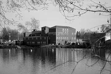 This piece features a black and white image of a building along the riverside in Baldwinsville New York that used to be the Red Mill Inn. The image is framed by branches from trees on the closest side of the river. In the distance , the building is flanked by smaller buildings and cars that lead into town on the left and Papermill Island on the right. The reflection of the building can be seen in the water but is distracted by the ripples in the water. 
