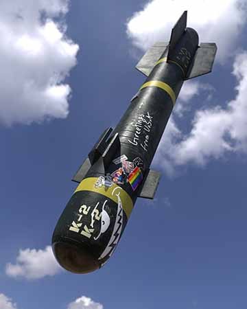 A commonly used tubular missile known as the hellfire missile is in the air with “woke” stickers and chalk drawings usually made by soldiers. There are some clouds in the background and the missile and the stickers are slightly worn out.