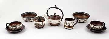 This ceramic tea set consists of two small plates, two handled cups, two bowls, one sugar jar, one creamer cup, and a teapot. Each is decorated with deep brown outlines of italian architecture, food, drink, and text on a white background. The rest of the details were glazed in a dark metallic silver and rustic and reddish brown.